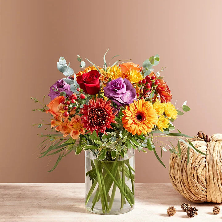 Colorful bouquet of flowers in a glass vase with a pumpkin and pinecones on a wooden surface.