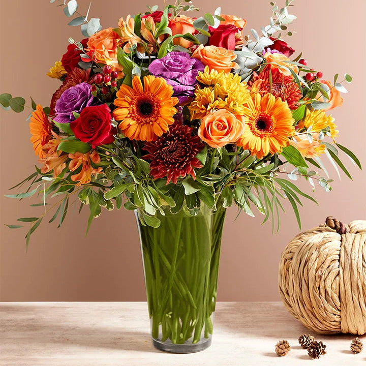Colorful bouquet of flowers in a glass vase with a pumpkin and pinecones on a wooden surface.