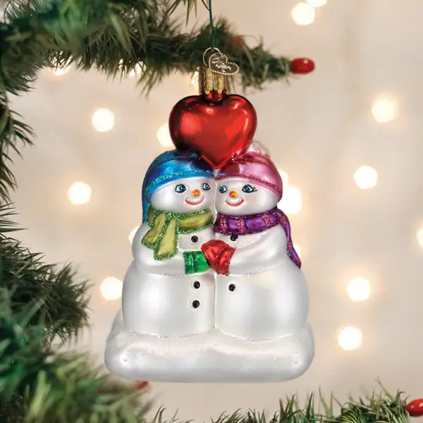 Christmas ornament of two snowpeople with a heart, hanging on a tree with lights in the background.