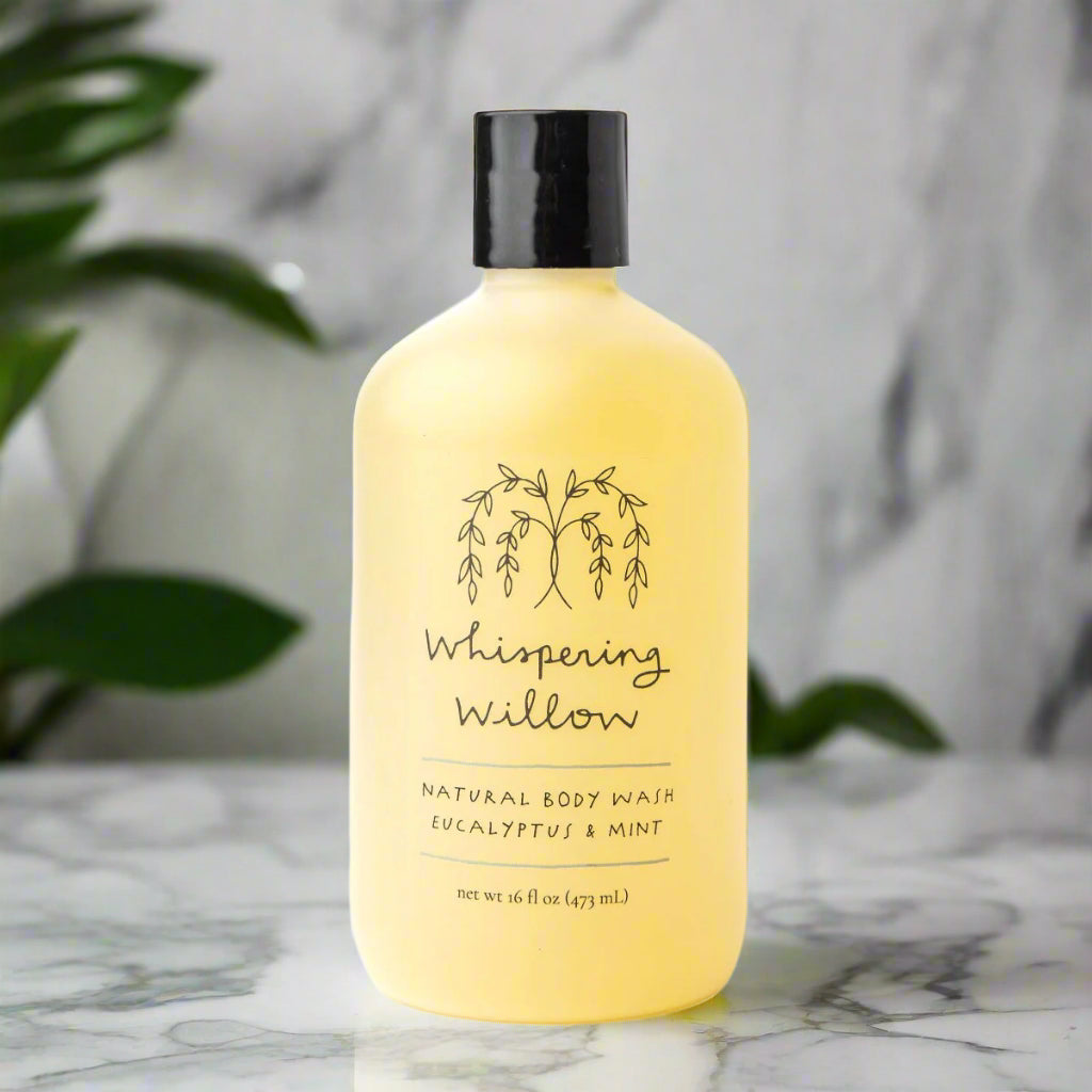 Yellow bottle of Whispering Willow natural body wash on a marble surface with green leaves in the background