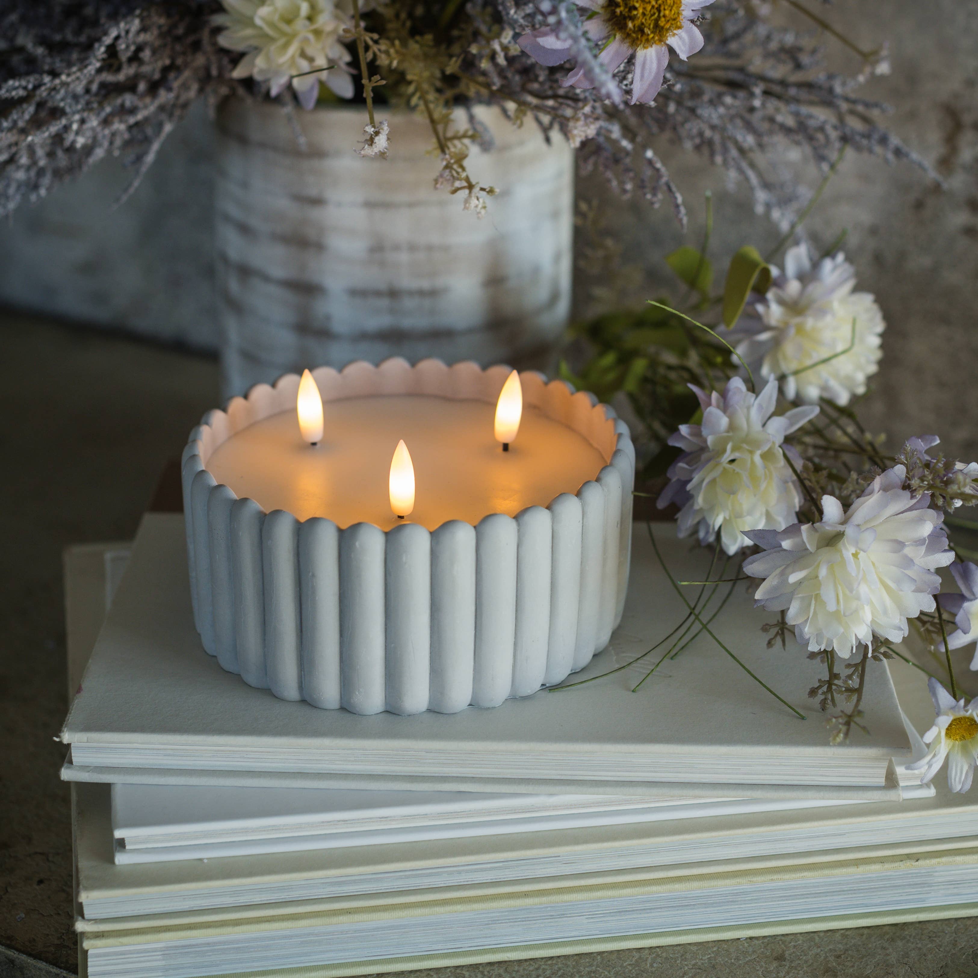 Ceramic candle holder with three lit candles on a stack of books, surrounded by flowers.