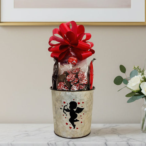 Gift basket with a large red bow in a metal bucket on a wooden surface.