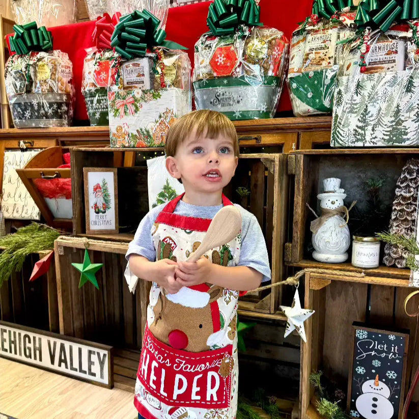 Child wearing a Christmas-themed apron with a wooden spoon in front of a display of gift bags and decorations.
