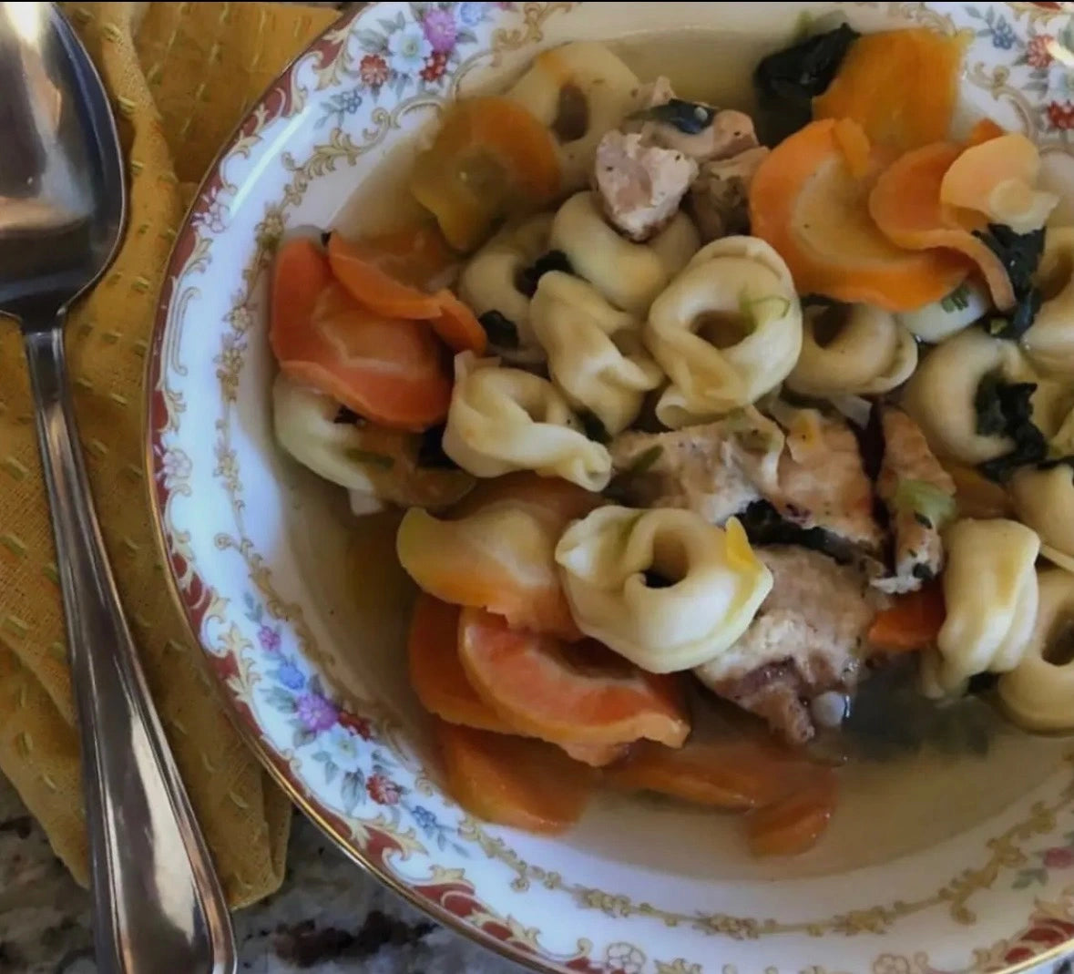 Bowl of pasta with carrots and chicken on a decorative plate with a spoon beside it.