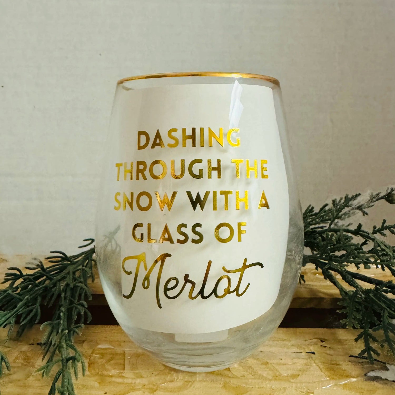 Glass with gold text 'Dashing through the snow with a glass of Merlot' on a wooden surface with greenery.