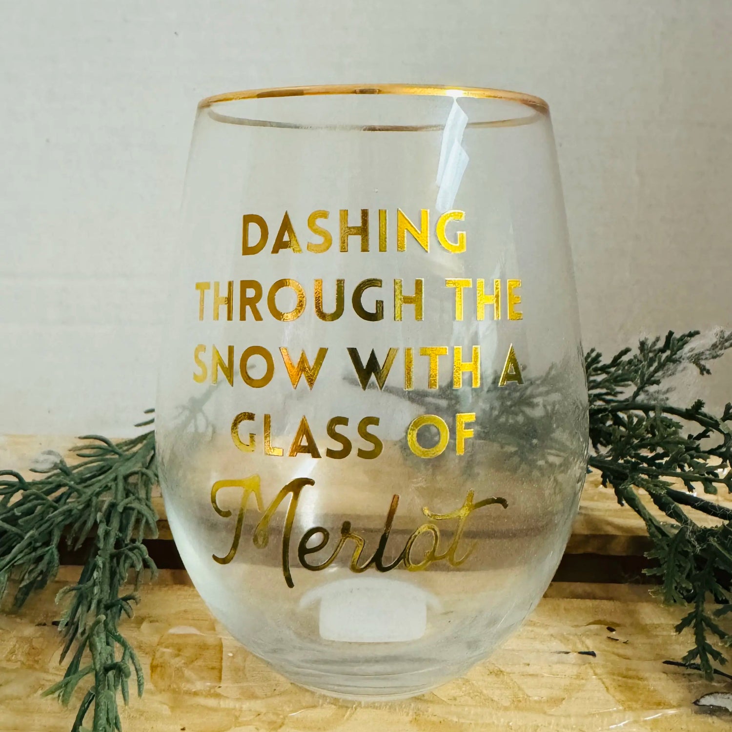 Glass with gold text 'Dashing through the snow with a glass of Merlot' on a wooden surface with greenery.