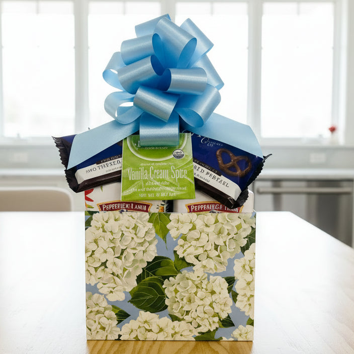 Gift basket with a blue bow, featuring vanilla cream spice tea and pretzels, on a wooden surface.