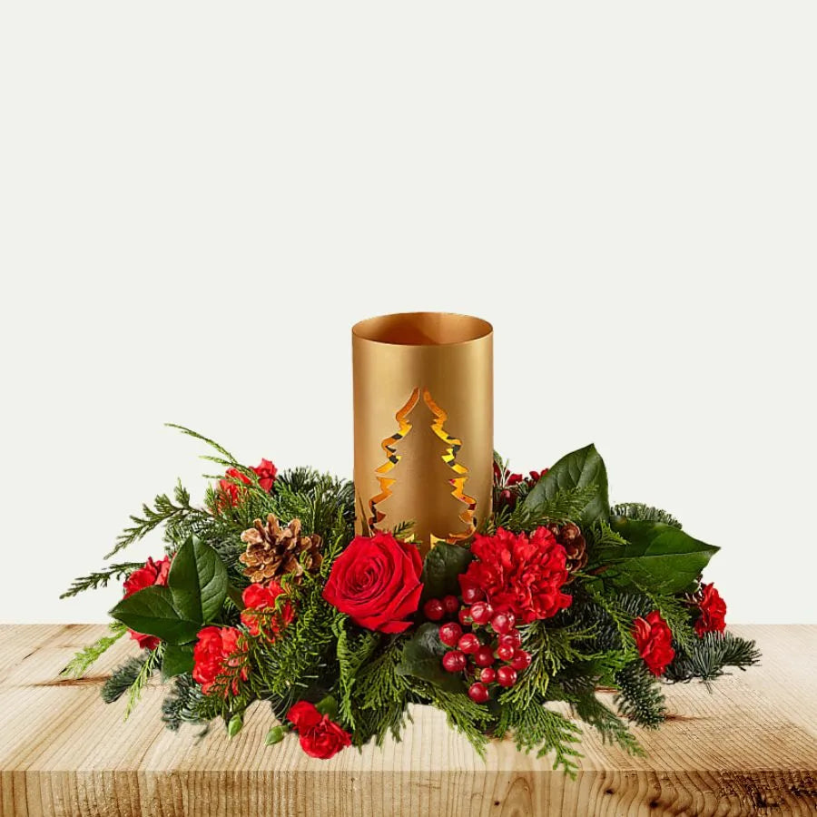 Decorative arrangement with red flowers, greenery, and a gold candle holder on a wooden surface.