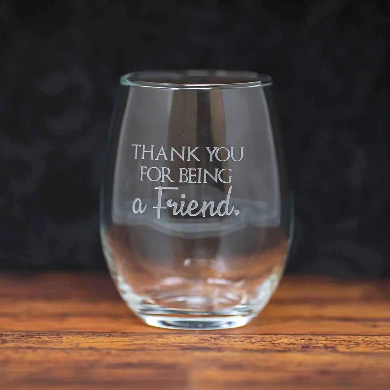Clear glass with 'Thank you for being a friend' text on a wooden surface with dark background