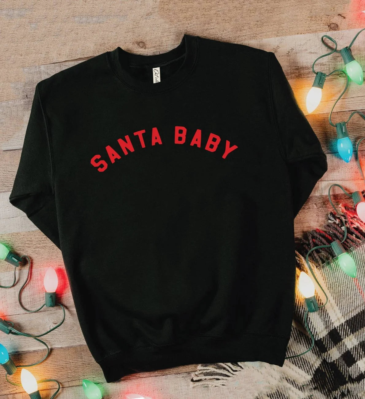 Black t-shirt with 'SANTA BABY' in red text on a wooden surface with Christmas lights.